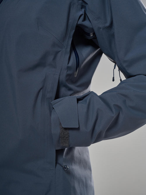Close-up of a dark sky blue waterproof jacket with a zippered pocket and adjustable cuff, shown on a neutral gray background.
