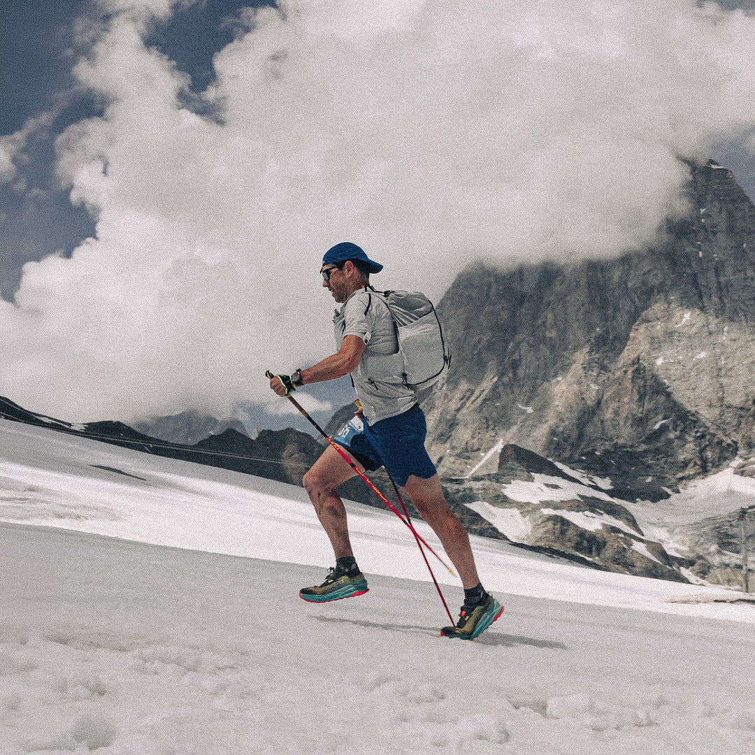 A male trail runner wearing a light grey t-shirt, blue shorts, and a blue cap is running up a mountain slope with a large rocky peak and dramatic clouds in the background.
