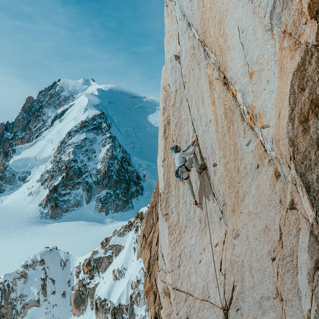 A rock climber scales a steep beige cliff face next to a snow-covered mountain under a blue sky.