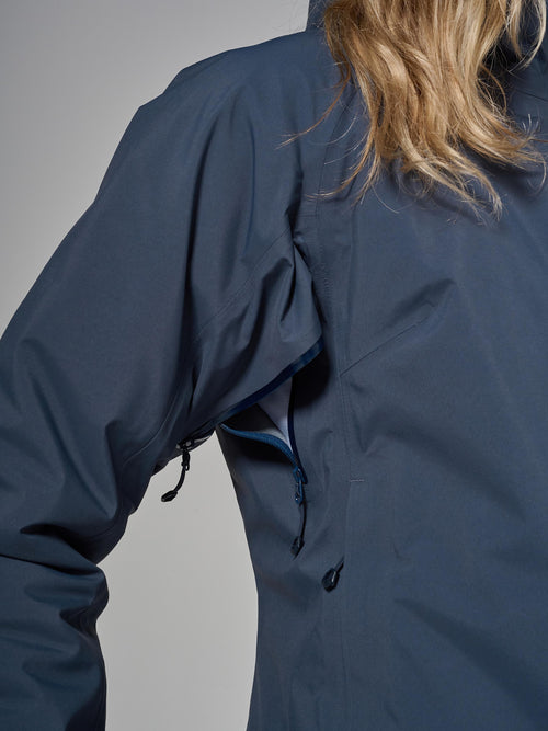 Close-up of a person wearing a dark sky blue waterproof jacket, showing pit zip and shoulder area against a neutral gray background.
