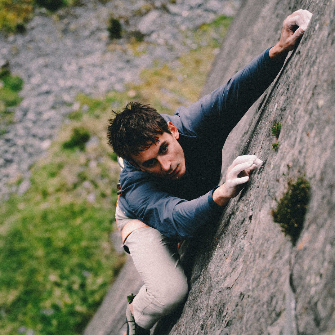 A man in a blue long-sleeve shirt and beige pants climbing a steep rock face, gripping holds with both hands and looking down with a focused expression.