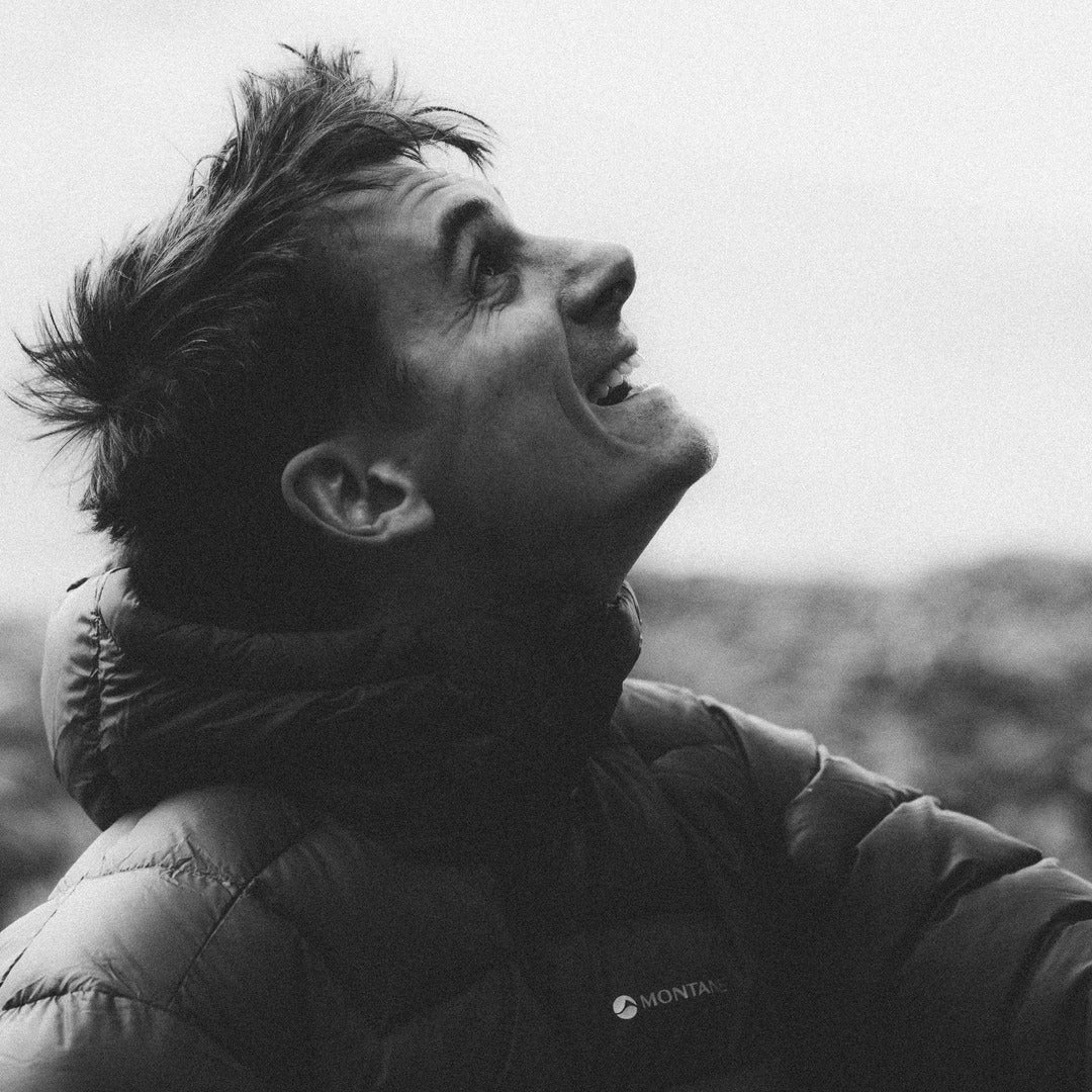 A black and white close-up photo of a smiling man wearing a puffer jacket with the brand name MONTANE visible on the chest, set against a soft, blurred outdoor background.