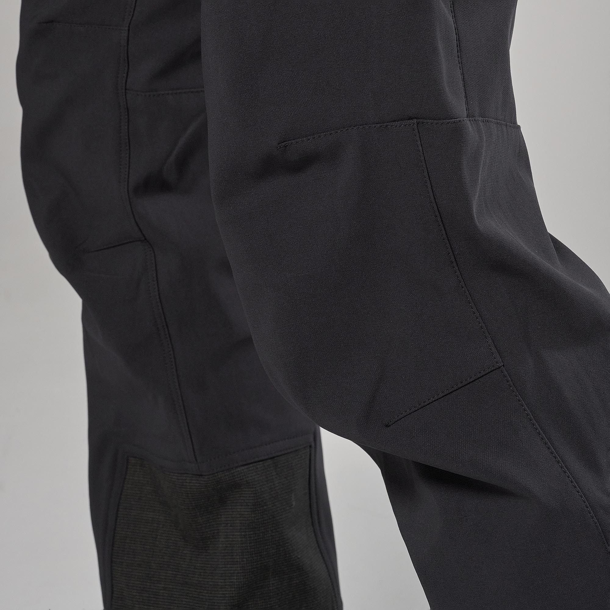 Close-up of dark gray technical pants showing the knee panel, seam details, and fabric texture against a neutral gray background.