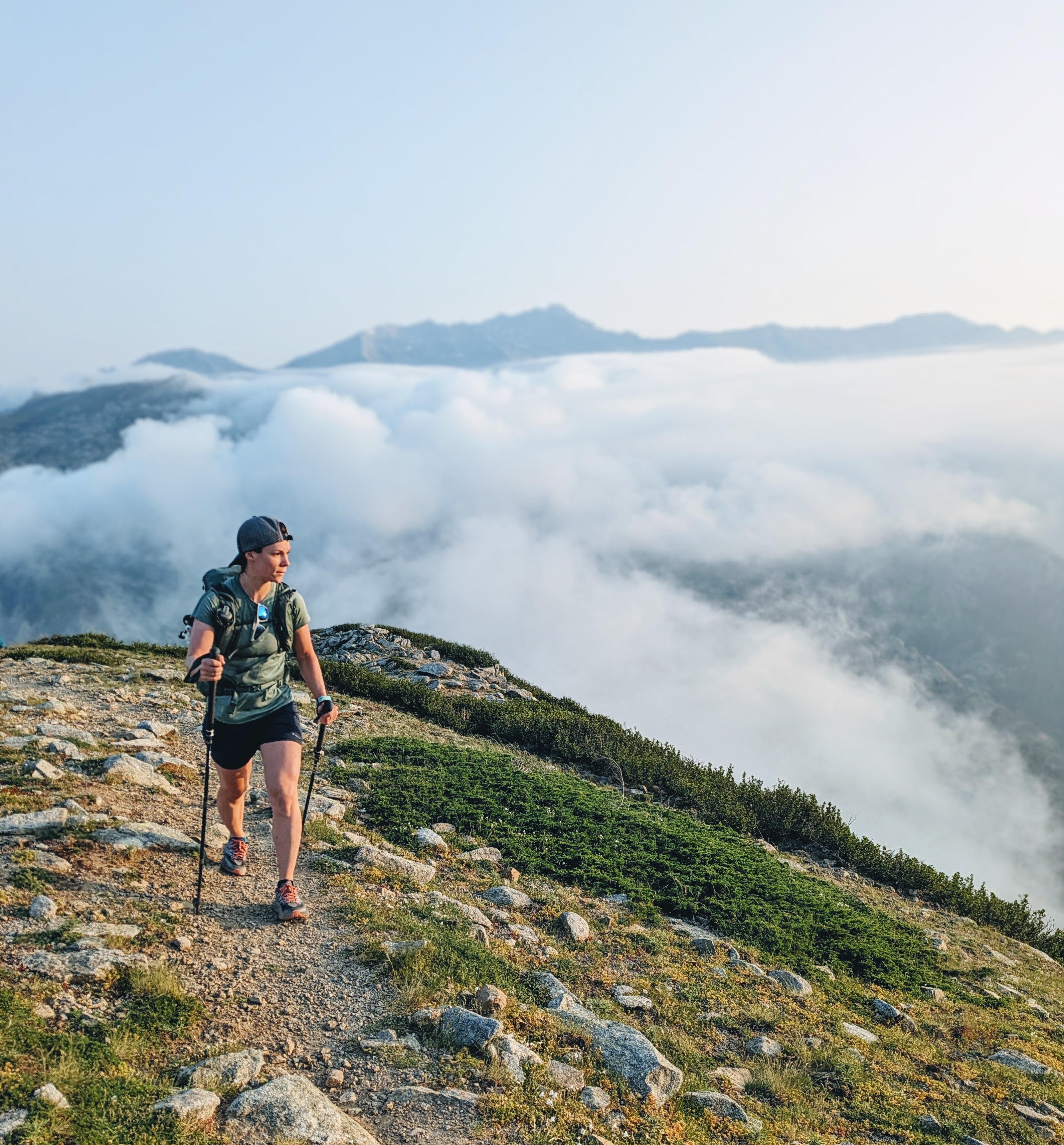 A hiker wearing a green camouflage t-shirt, black shorts, and a backpack walks along a rocky trail on a mountain ridge with a sea of clouds below.